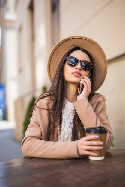 Sognando la modella modella è seduta sul tavolo di abiti da caffè in abiti casual occhiali da sole scuri con una tazza di caffè