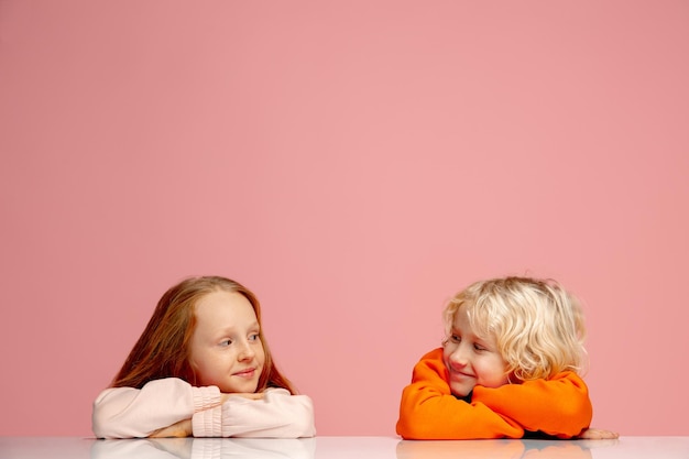 Dreams. Happy children isolated on coral pink studio background. Look happy, cheerful. Copyspace for ad. Childhood, education, emotions, facial expression concept. Sitting at the table dreamful.