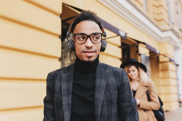 Dreamy african man in glasses standing on the street. Outdoor photo of stylish black guy listening music in headphones.