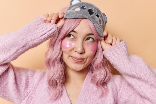 무료 사진 분홍색 머리를 한 꿈꾸는 아시아 여성은 부드러운 수면마스크를 쓰고 캐주얼한 점퍼는 눈 밑에 미용 패치를 적용하여 붓기를 제거하거나 주름이 갈색 배경 위에 신중하게 고립된 모습을 보입니다