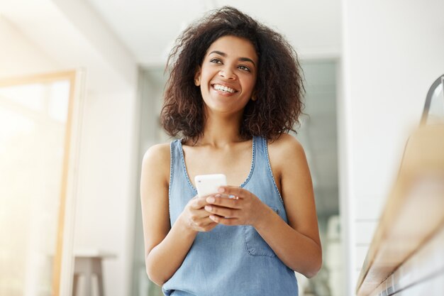 夢のような美しいアフリカ女性がカフェに座っている携帯電話を保持している夢を見て考えを笑っています。