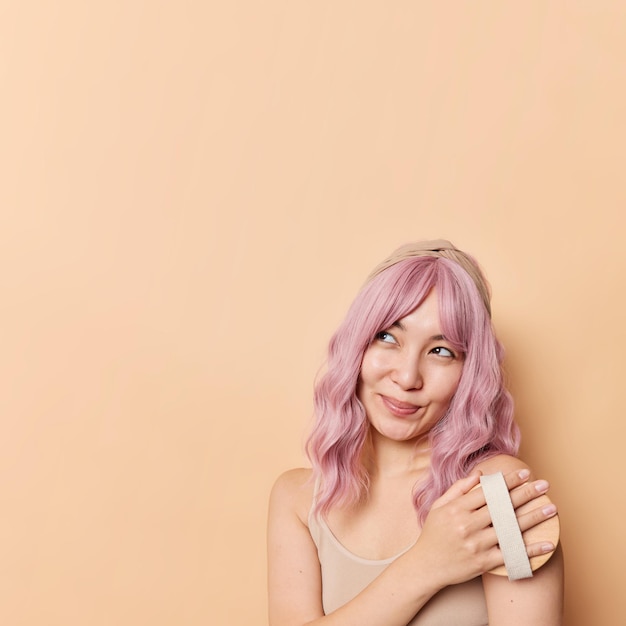 Dreamy beautiful Asian woman with dyed pink hair brushes body with dry brush has healthy smooth skin looks away smiles gently poses against beige background blank copy space for your promotion