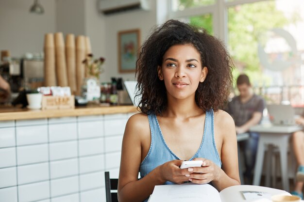 Dreamy beautiful woman student sitting in cafe with books and magazines smiling holding phone thinking.