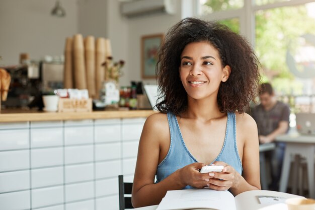 夢のような美しい女性学生の本や雑誌を保持している電話の思考を笑顔でカフェに座っています。