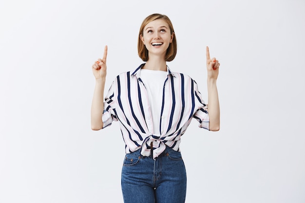 Free photo dreamy beautiful young woman pointing fingers up and looking amazed