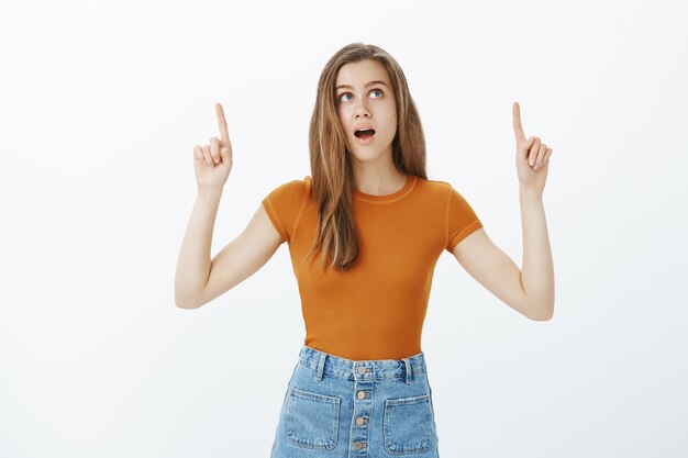 Dreamy beautiful young woman pointing fingers up and looking thoughtful, making choice, smiling while deciding