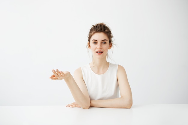 Dreamy beautiful young woman with bun sitting at table over white background thinking dreaming