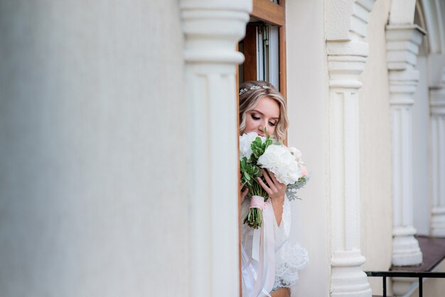 Dreamy bride with wedding bouquet stands behind the column in the hall