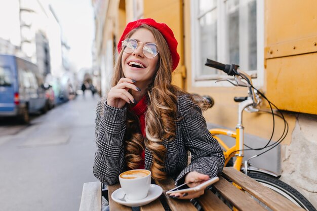 通りでコーヒーを飲みながら笑っている夢のような茶色の髪の少女。ラテと自転車のカップを背景に木製のテーブルに座っている赤いベレー帽のロマンチックな女性の肖像画。