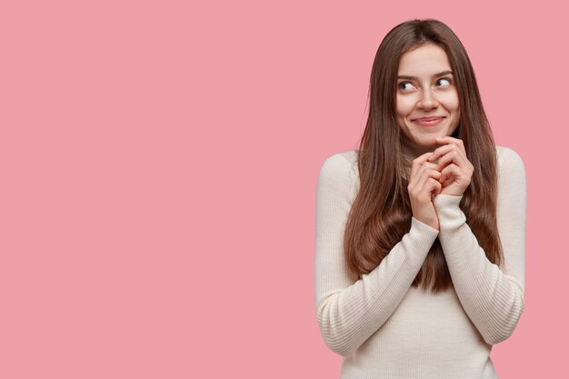 Dreamy brunette woman smiles gently, keeps hands together under chin, wears casual clothes