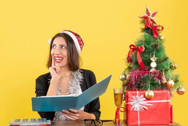 Мечтательная бизнес-леди в костюме в шляпе санта-клауса и новогодних украшениях проверяет документ и сидит за столом с рождественской елкой в офисе