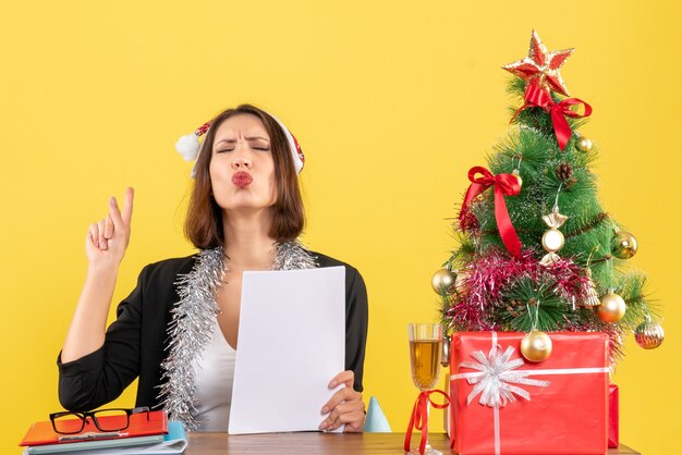 산타 클로스 모자와 새해 장식 양복에 꿈꾸는 비즈니스 아가씨는 혼자 위를 가리키고 사무실에서 크리스마스 트리가있는 테이블에 앉아 있습니다.