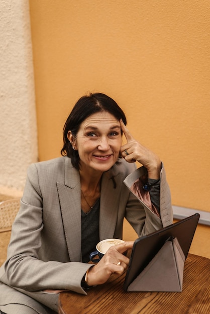 Мечтательная кавказская взрослая брюнетка улыбается, работая на планшете и пьет кофе в уличном кафе Концепция бизнес-работы образования
