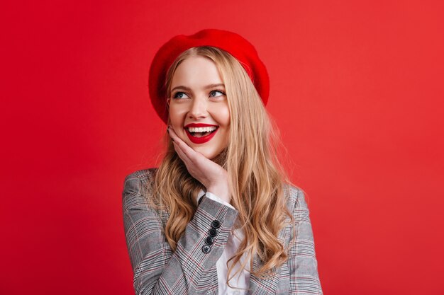 Dreamy caucasian girl with blonde hair looking up with smile.  positive french woman in beret isolated on red wall.