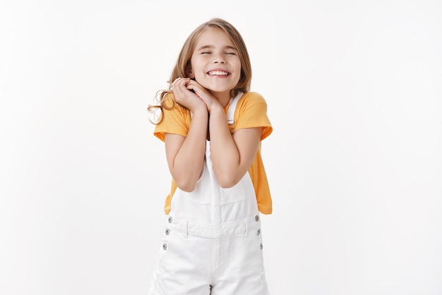 Dreamy cheerful happy little girl with blond hair, clasp hands together joyfully cannot wait outstanding event, close eyes smiling dreamy, feeling overjoy and happiness, stand white wall