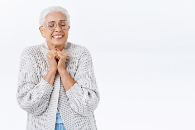 Dreamy, cheerful old woman feeling affection and joy as remember nice memory, press hands together near chest, close eyes and smiling excited, pleased kids come home for holidays