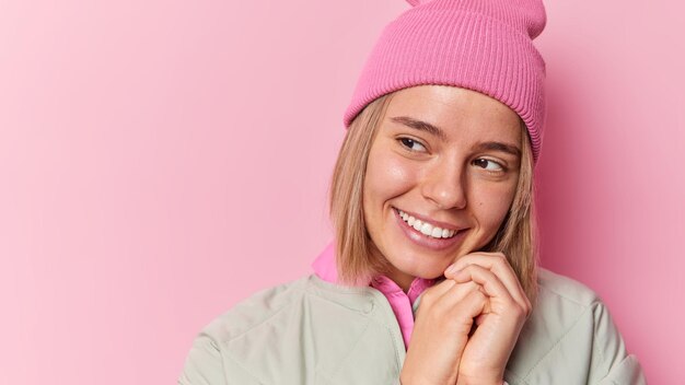 Dreamy cheerful woman keeps hands near face smiles gently looks away thinks and recalls about something pleasant wears hat and jacket isolated over pink background blank copy space for promo