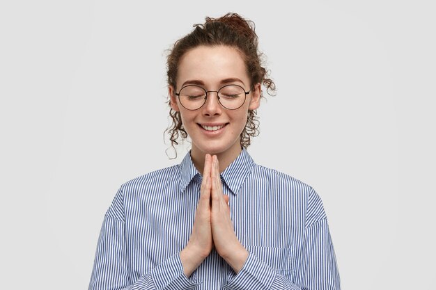 Dreamy cheerful young female keeps plams pressed together, asks God for successful personal life and family, has curly hair, freckled skin, stands alone against white wall, enjoys calm atmosphere