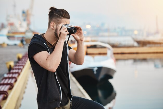 Dreamy creative european photographer in stylish outfit standing in harbour, taking picture of sea