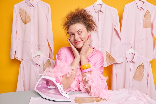 Dreamy curly haired housewife poses near ironing board being deeps in thoughts daydreams while doing domestic work irons freshy washed clothes isolated over yellow wall. Maid tides up laundry