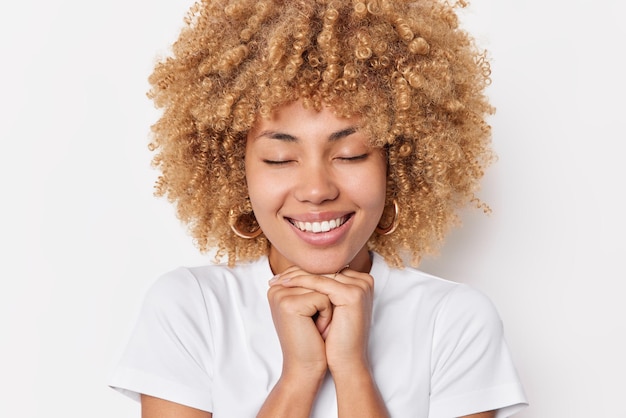 Dreamy curly haired young woman keeps hands under chin smiles pleasantly recalls something pleasant dressed in casual t shirt isolated over white background. Happy emotions and feelings concept