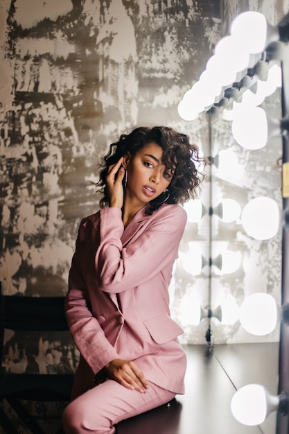 Dreamy curly woman in pink suit sitting near mirror