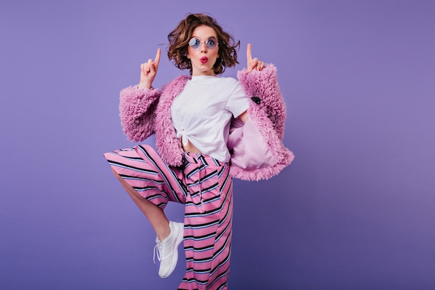 Dreamy dark-haired lady jumping on purple wall. Sensual girl in white sneakers and pink pants having fun at indoor photoshoot.