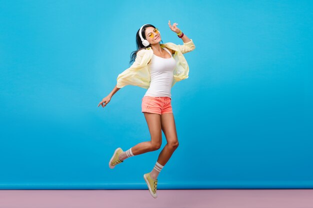 Dreamy dark-haired latin woman in casual colorful attire dancing. Indoor photo of romantic young lady with happy face expression jumping in room with blue walls.