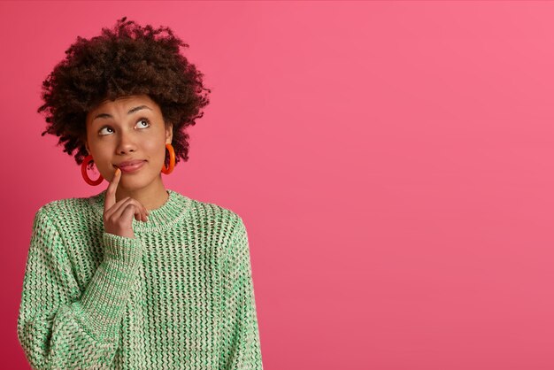 Dreamy dark skinned young woman thinks about career opportunities, stands thoughtful, concentrated upwards, dressed in knitted sweater, isolated on pink wall, copy space for your promotion