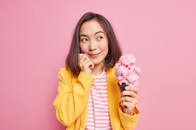 Мечтательная восточная женщина приятно смотрит в сторону с задумчивым выражением лица, держит большое вкусное мороженое, думает о чем-то, держит руку на лице, одетая в стильную одежду, изолированную на розовой стене