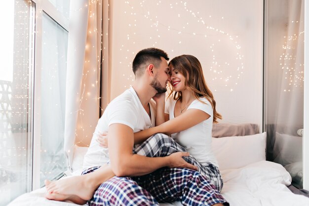 Dreamy european woman sitting on boyfriend's knees. Barefooted romantic girl chilling with husband in morning.