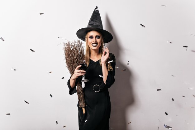 Dreamy evil witch drinking wine. Ecstatic young woman with blonde hair smiling at halloween party.