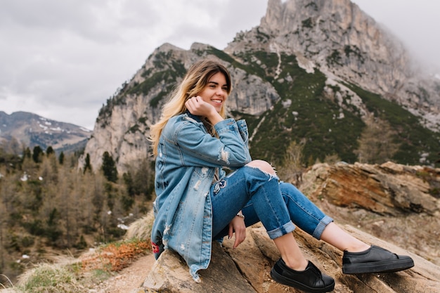 Dreamy fair-haired girl wearing denim attire resting on the rock after hard climbing and posing touching her face