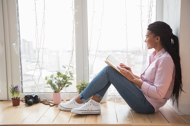 Dreamy female student with book sitting on window sill