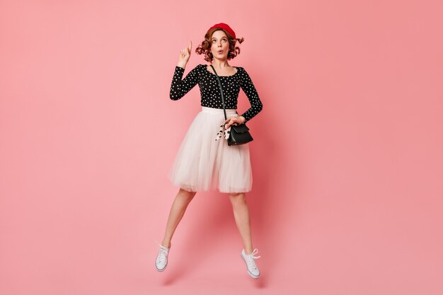 指で指している夢のようなフランスの女の子。ピンクの背景に身振りで示すベレー帽の素敵な女性のスタジオショット。