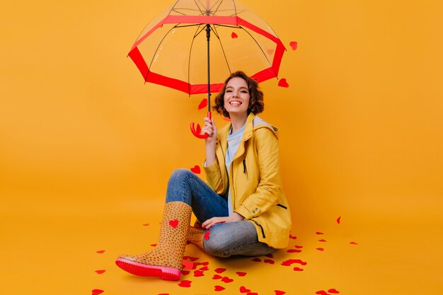 Мечтательная девушка в забавных резиновых туфлях, сидя со скрещенными ногами на желтой стене. Крытый портрет романтичной женщины в яркой куртке, позирующей с зонтиком.