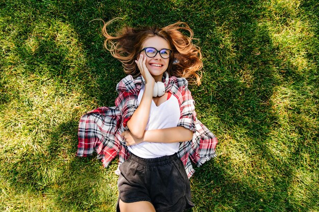 Dreamy girl in headphones lying on grass and looking at sky Positive smiling woman spending weekend morning in park