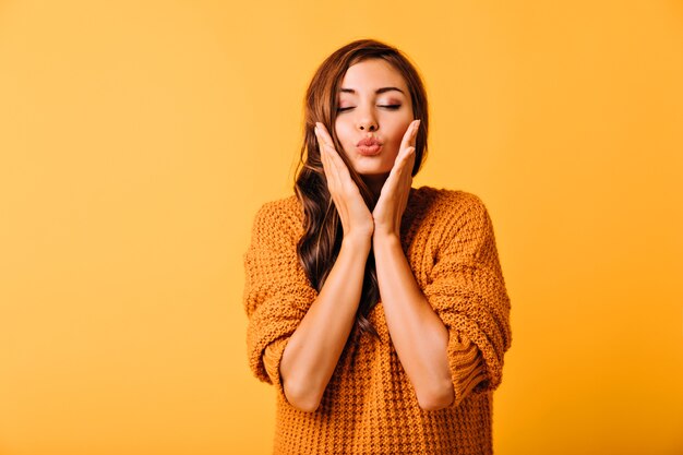 Dreamy girl in knitted clothes touching her face. Inspired brunette woman with light makeup standing on yellow.