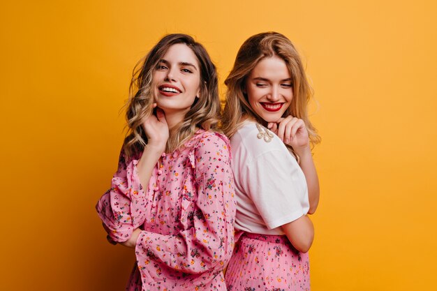 Dreamy girl in pink blouse posing with her sister. Lovable female friends laughing on yellow wall.