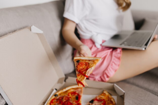 ソファに座ってピザのスライスを保持しているピンクのショートパンツの夢のような女の子。洗練された白人女性がコンピューターを食べて作業している屋内ショット。