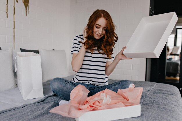 Dreamy girl in striped tshirt sitting on bed Ecstatic redhaired curly woman posing in bedroom