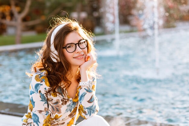 Dreamy girl in white wristwatch and glasses sitting on beautiful fountain propping face by hand