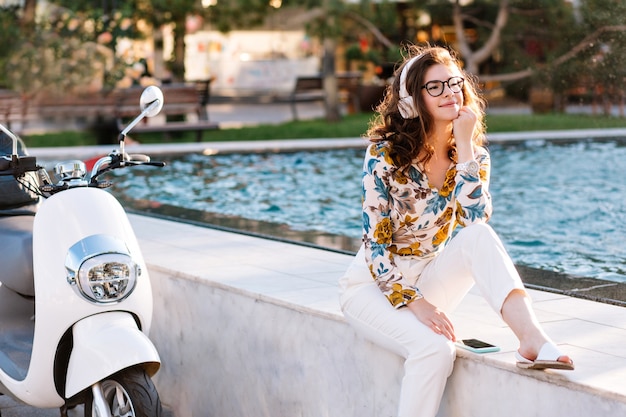 Dreamy girl with elegant hairstyle listening music and looking away, spending time near fountain