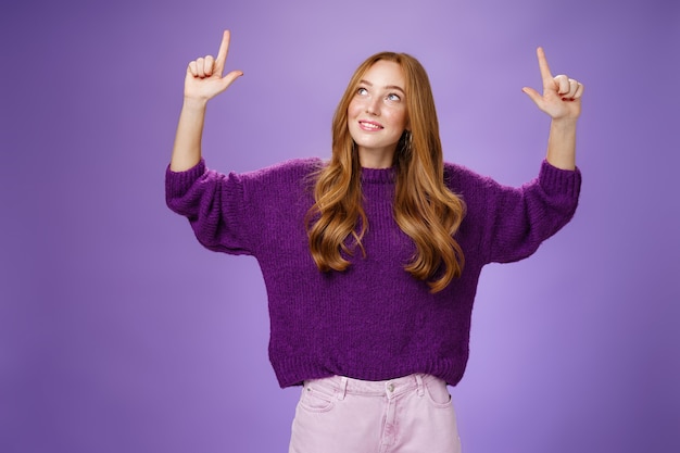Мечтательная девушка с рыжими волосами и веснушками в теплом уютном фиолетовом свитере поднимает руки, смотрит и указывает вверх с заинтригованным и счастливым очарованным выражением лица, улыбаясь, как любопытный продукт над фиолетовой стеной.