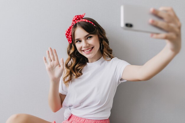 Мечтательная девушка с волнистой прической позирует с улыбкой. Снимок в помещении замечательной молодой леди в белой повседневной футболке, делающей селфи.