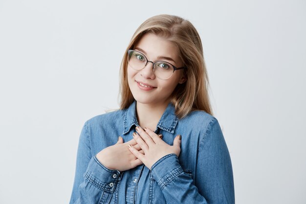 Dreamy glad female looks  with pleasure and gratitude, keeps hands together on chest, smiles happily, imagines pleasant moments in her life. Young blonde woman in denim shirt and eyewear