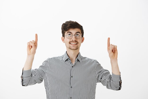 Dreamy good-looking ordinary man in round glasses, smiling curiously while looking and pointing up with index fingers