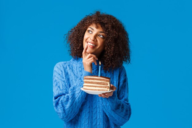 Dreamy and happy, beautiful african-american woman with afro haircut, thoughtful gazing up, smiling and touching her lip as if thinking what wish for before blowing-out candle in birthday cake.