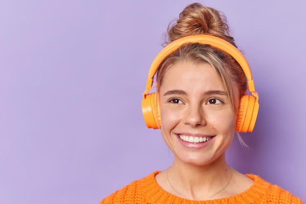 Free photo dreamy lovely young woman smiles broadly enjoys audio track in wireless headphones concentrated away with dreamy expression isolated over purple background blank empty space for your promotion