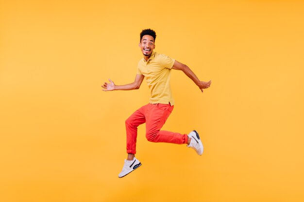 Dreamy male model with brown skin fooling around. Indoor shot of african guy in bright clothes jumping.